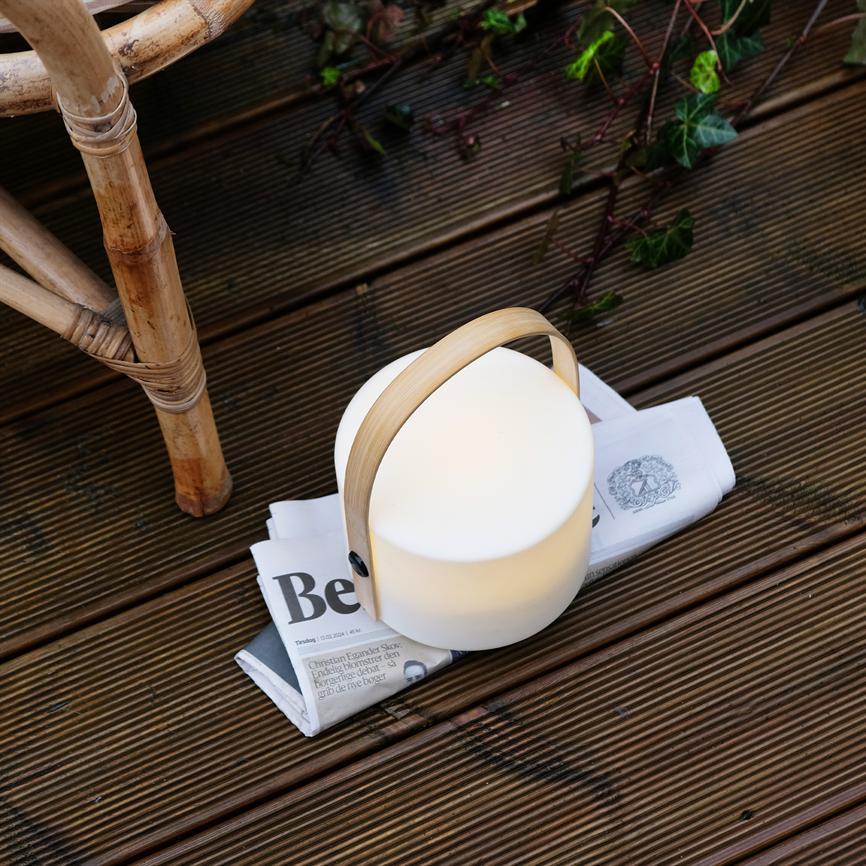Round portable lamp with bamboo handle emits soft light, stands on a folded newspaper on a dark wooden terrace by a rattan chair and climbing ivy. Text: "Pray"
