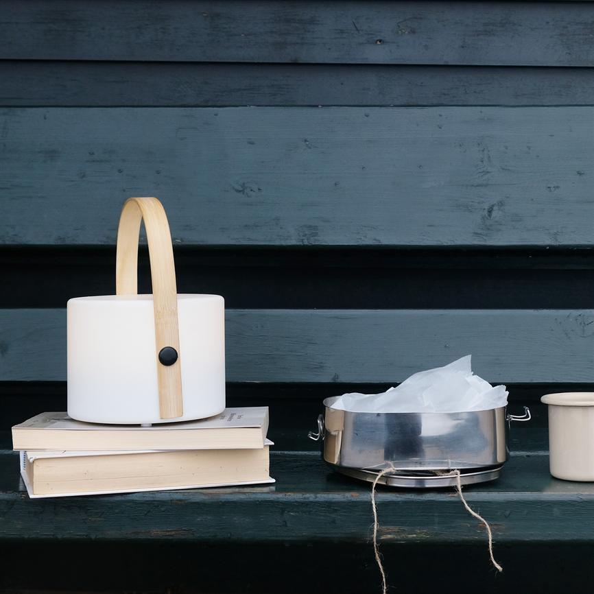 Lantern with cylindrical white shade and wooden handle stands on two stacked books; next to a metal container with parchment paper and a ceramic mug on a dark bench in front of a blue wooden wall.