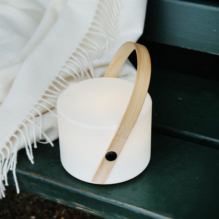 A portable white lamp with a curved wooden handle shines softly on a dark green wooden bench next to a white fringed rug.