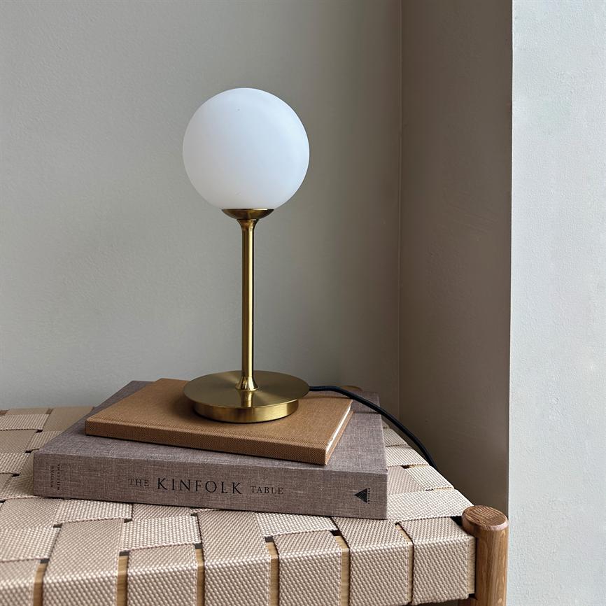 Table lamp with round white glass shade and brass stand stands on two books on a wicker bench in a bright corner of the wall. 
 Text on spine: "THE KINFOLK TABLE".