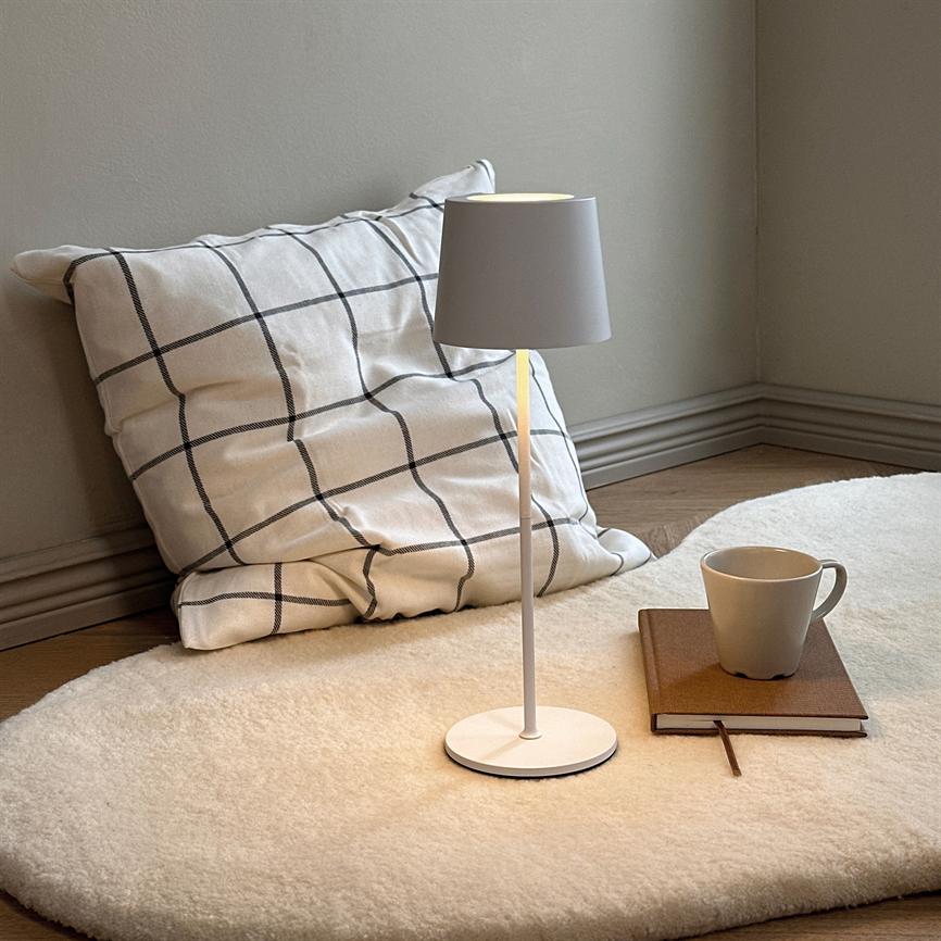 Table lamp is lit on a round, bright woolen mat; next to it is a checkered pillow leaning against the wall and a notebook with a mug on the wooden floor in a quiet corner.