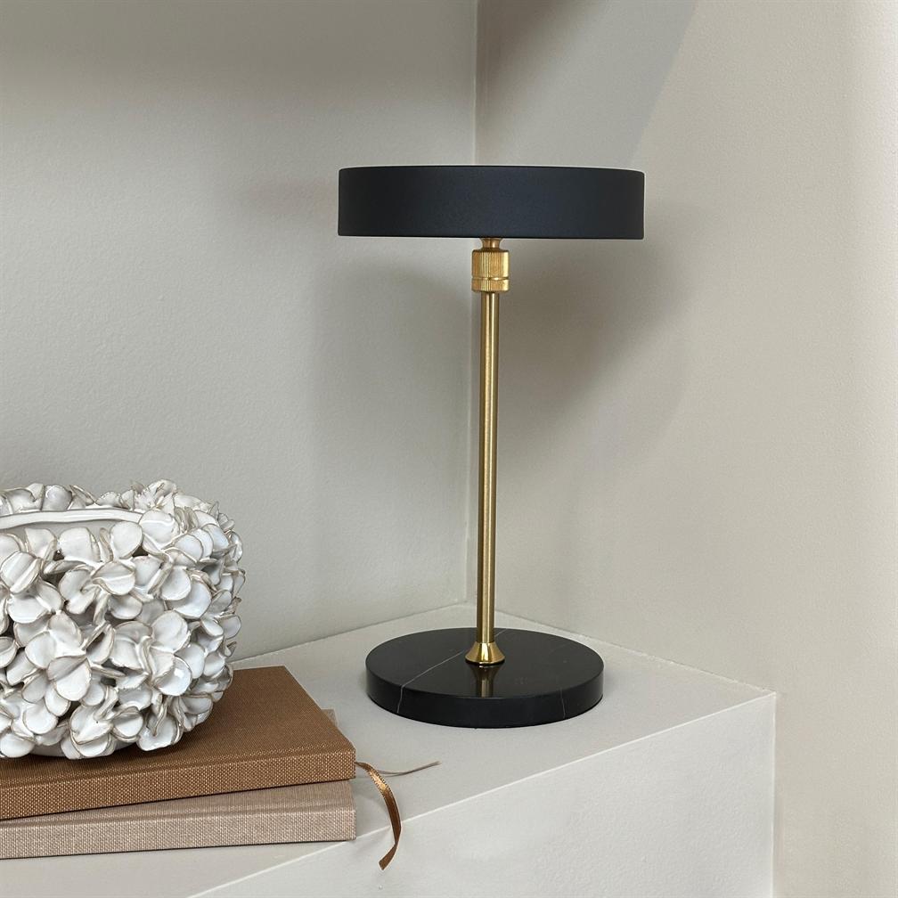 Table lamp with black shade and gilded rod stands on a white corner table; next to it are two cloth-bound books and a white decorative bowl shaped like flowers.