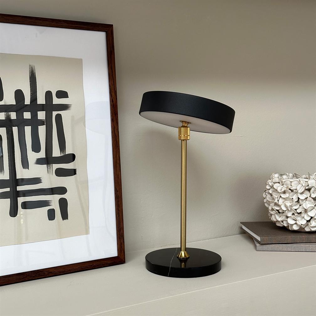 En lille bordlampe med sort rund skærm og messingstang står med skærmen let tiltet på en hylde ved en neutral væg, flankeret af indrammet abstrakt tryk, bøger og en hvid skulptur.