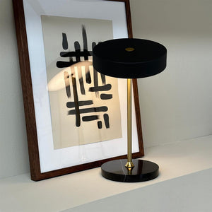 Table lamp is lit on a white shelf; it has a black round shade, slim gold bar and black marble base; the light casts a soft glow on the framed abstract black-and-white print behind.