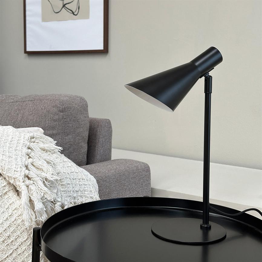 Black metal table lamp leans over a round black tray table; it stands next to a gray sofa with a white braided rug and a neutral wall in the background.