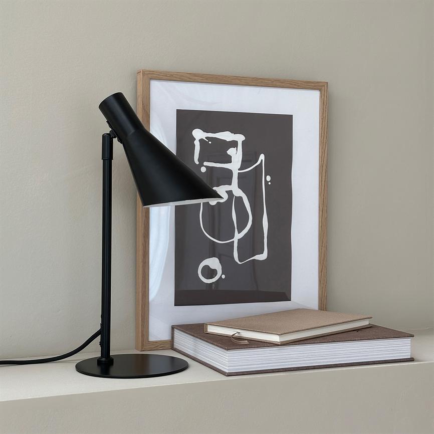Desk lamp is tilted forward and illuminates a framed abstract poster; black lamp and wooden frame stand on a shelf with two books in front of a beige wall.