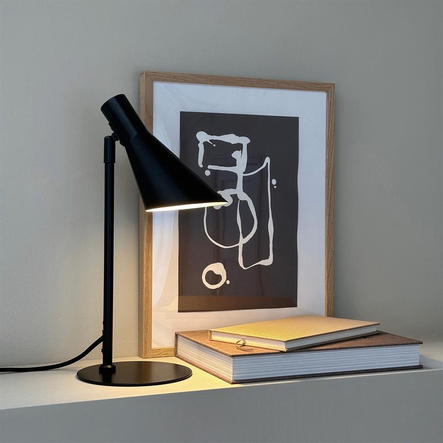 A black table lamp shines down on a stack of books and a framed abstract motif. The objects stand on a white shelf in front of a neutral gray wall.
