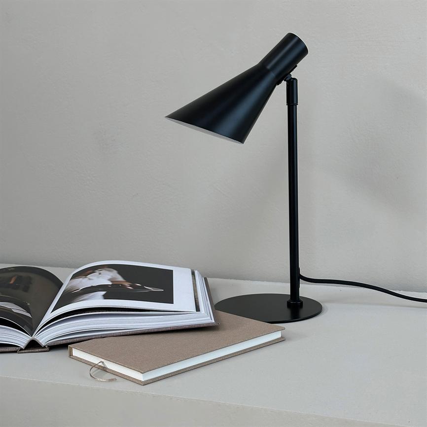 Black metal table lamp points down over an open photo book; a closed notebook lies next to it on a light table top with a visible cord in front of a light gray wall.