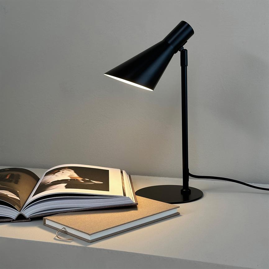 A black desk lamp shines on an open photo book and a linen-bound notebook on a bright table in front of a plain gray wall.