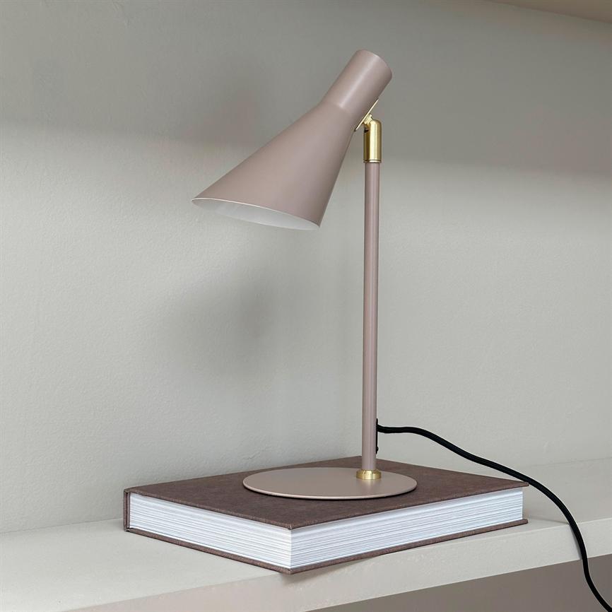 Table lamp points obliquely downwards, stands on a closed, thick book on a shelf in front of a light gray wall; metal rod and shade in dusty pink with black cord.