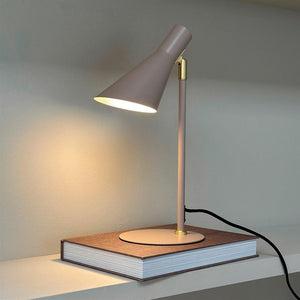 Table lamp shines softly on a thick bound book, placed on a shelf in front of a light wall; slim dusty pink lamp with gold detail and visible black cord.
