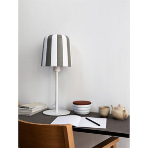 Table lamp with vertical white and gray stripes stands on a desk; open notebook and pen in front, cup, teapot and bowl beside; wooden chair in the foreground, white wall behind.