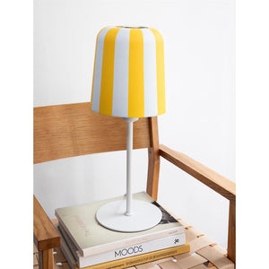 A yellow and white striped table lamp sits on a stack of books on the seat/armrest of a wooden chair in a bright, minimalist room. Text on books: "MOODS"; "BUILDING CULTURE AND CONSERVATION VALUES".