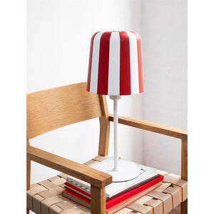 Table lamp with red and white striped shade stands on a stack of books on the seat of a wicker wooden chair in a bright, minimalist room.