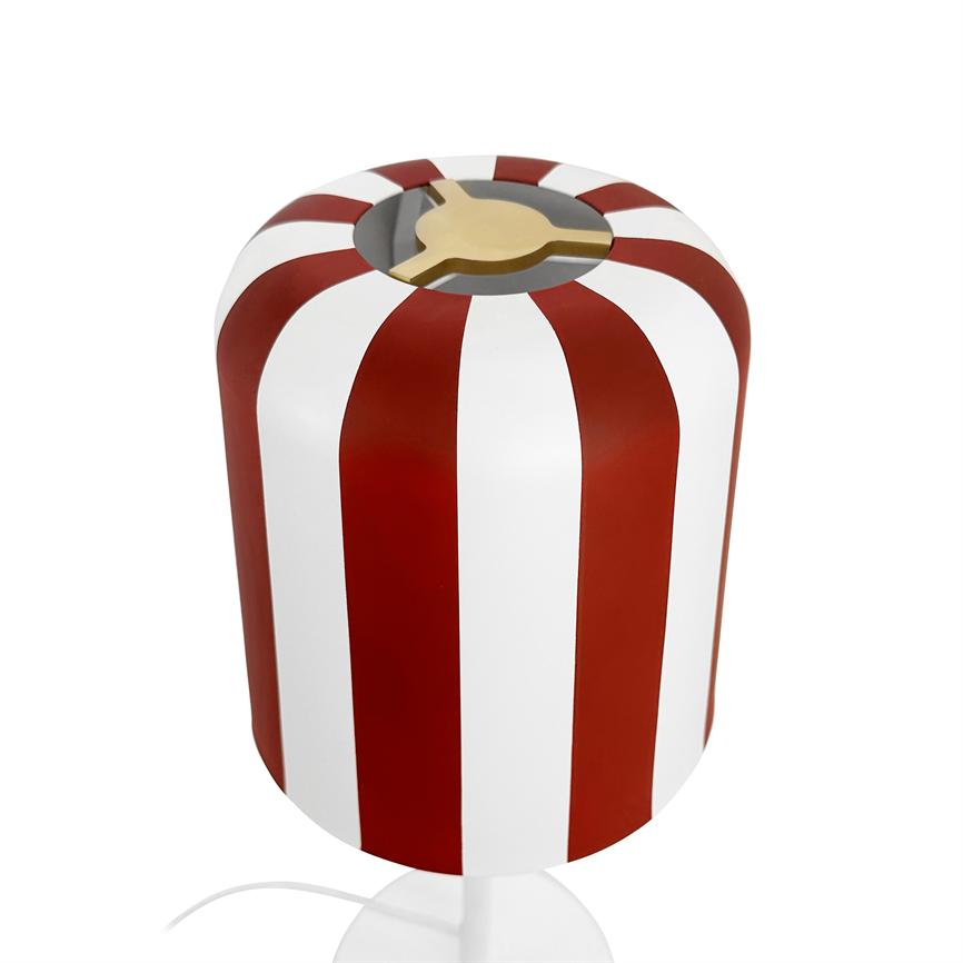 Cylindrical table lamp with red and white vertical stripes, golden three-arm inlet at top. Standing on white surface; white wire seen from bottom, isolated white background.