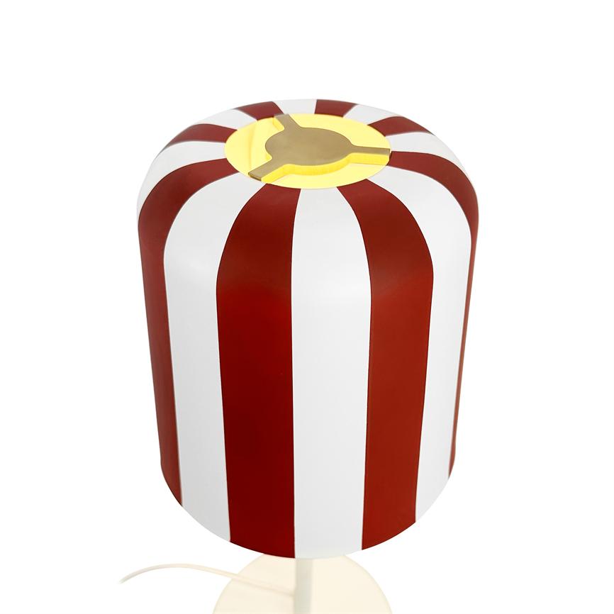 Cylindrical lampshade with red and white vertical stripes emitting a warm yellow light from the top; stands on a light background with visible wire.