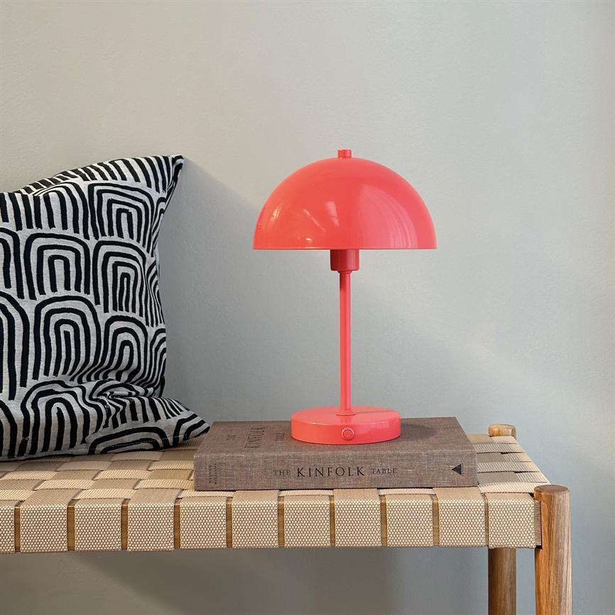 Coral red table lamp stands on a book (“THE KINFOLK TABLE”) on a wicker bench; black and white, geometrically patterned pillow at left; light, neutral wall in the background.
 
 Text: THE KINFOLK TABLE