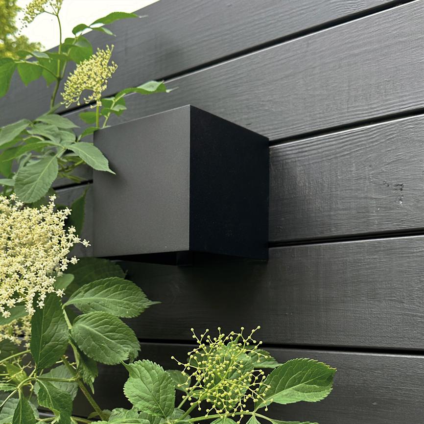 Black cube-shaped outdoor lamp mounted on a dark wooden fence, surrounded by green leaves and small white flowers; modern, minimalist expression in a garden context.