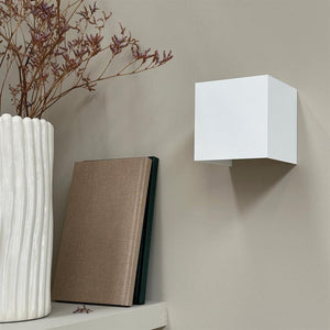 White cubic wall lamp mounted on a beige wall, casts a shadow below; to the left a shelf with a tall white vase of dried flowers and two books standing against the wall.