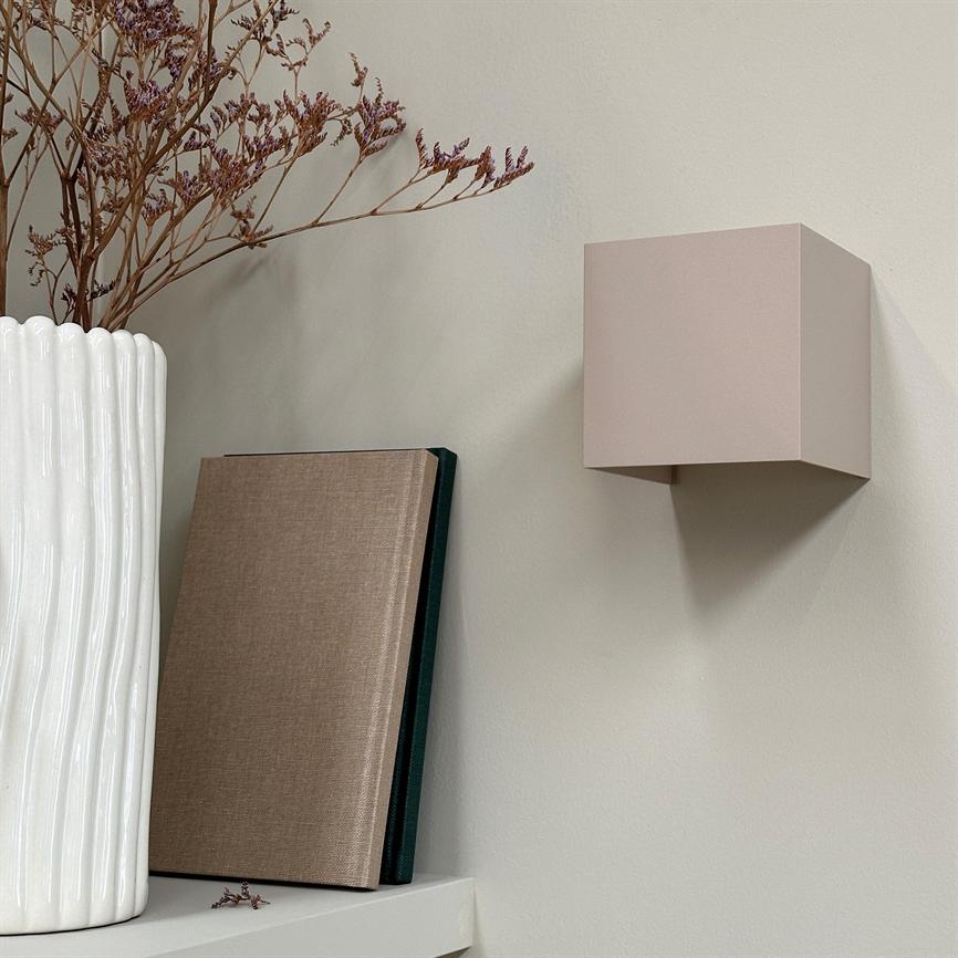 A cubic wall lamp hangs and casts a shadow on a light wall; on a nearby shelf is a white fluted vase with dried flowers and two bound books.