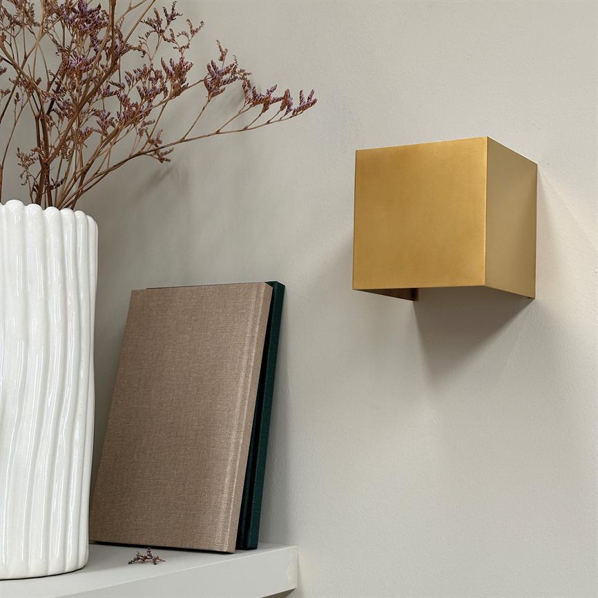 Gold colored cube wall lamp hangs on a light wall and casts a soft shadow; next to it is a white ceramic jar with dried flowers and two bound books on a shelf.