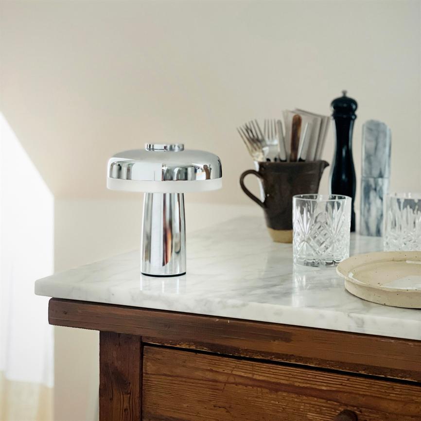 A chrome table lamp stands on a white marble top; in the background a jar with cutlery, a pepper grinder, crystal glass and a round plate on a wooden cabinet.