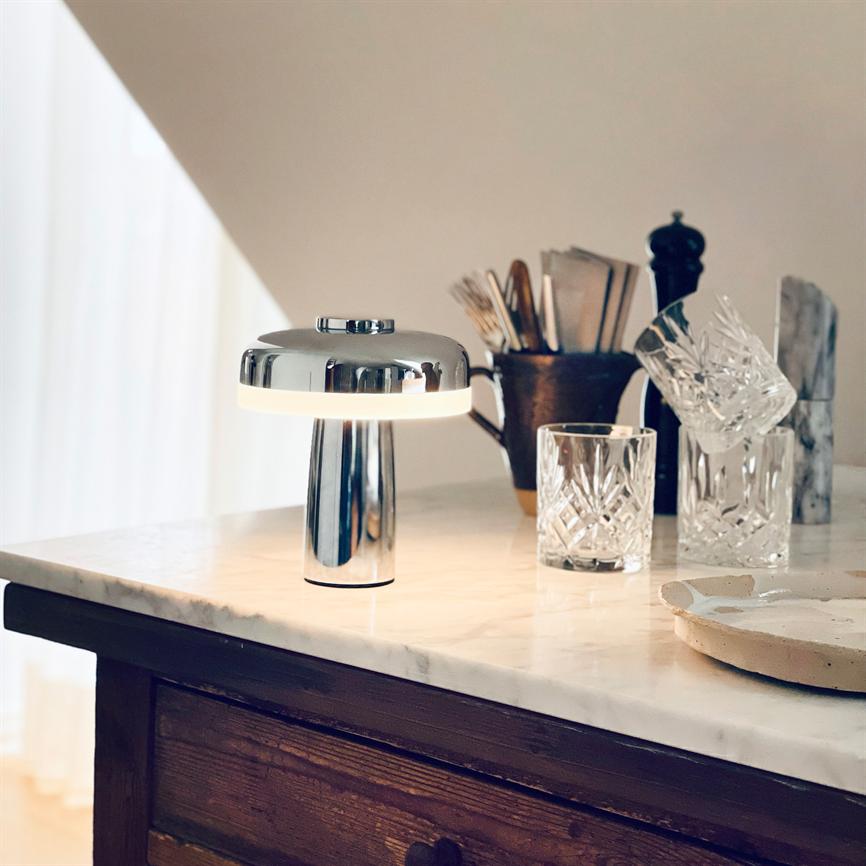 A chrome table lamp casts soft light on a marble tabletop, surrounded by crystal glass, a cutlery mug and a serving dish on a dark wooden cabinet in a bright kitchen corner.