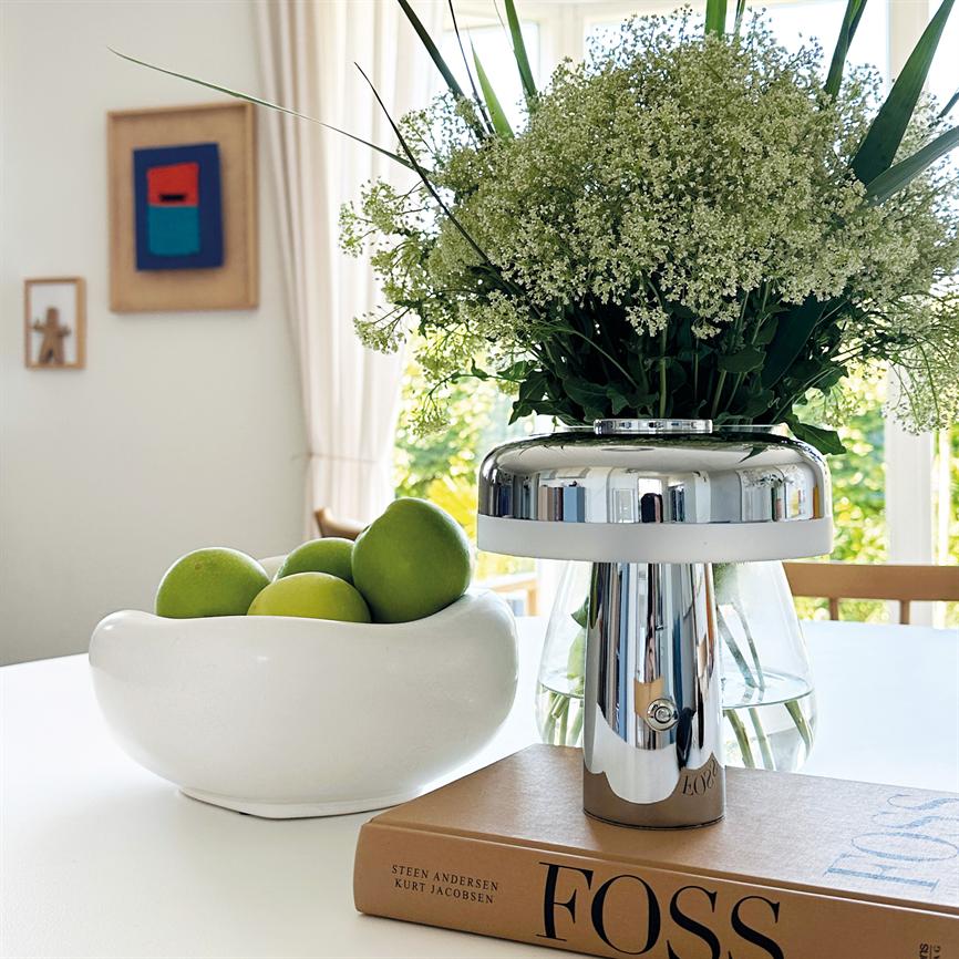 A chrome table lamp stands on a book on a bright table and mirrors the room; next to a white bowl with green apples and a glass vase with white flowers. Text: "FOSS"; "STEEN ANDERSEN KURT JACOBSEN".