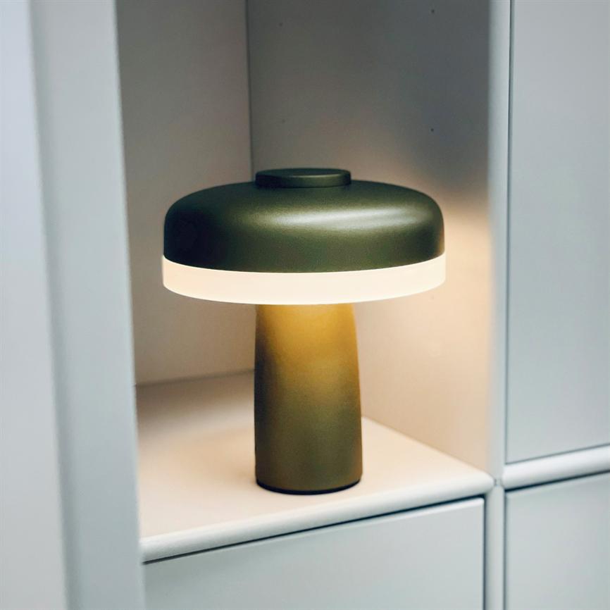 An olive green table lamp shines softly from its white diffuser, standing on a shelf in a white built-in bookcase, creating a warm, subdued light in the niche.