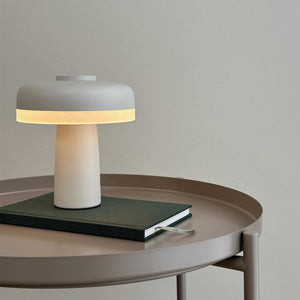 Lamp emits soft, warm light; stands on a green bound notebook on a round beige metal table in front of a light gray wall.