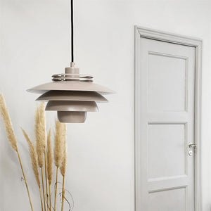 A layered, beige pendant lamp hangs on a black cord, in the middle of a white wall; pampas grass stands to the left and a closed paneled door is seen to the right.