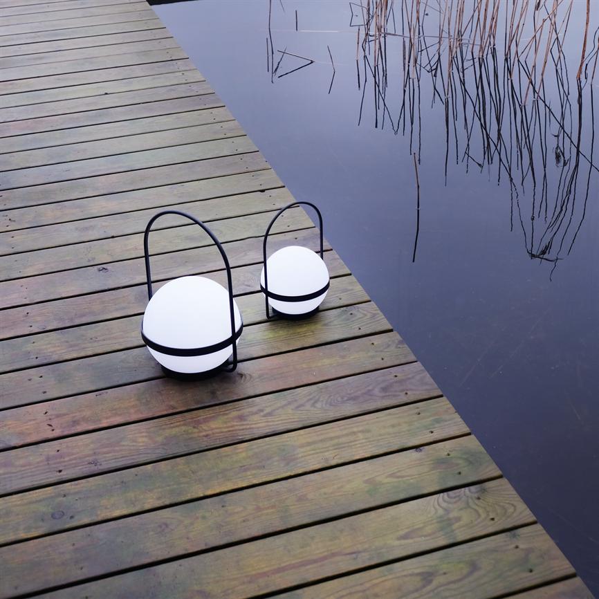 Two round lanterns rest on a wooden bridge, white balls in black hoops, by still dark water with reeds and reflections.