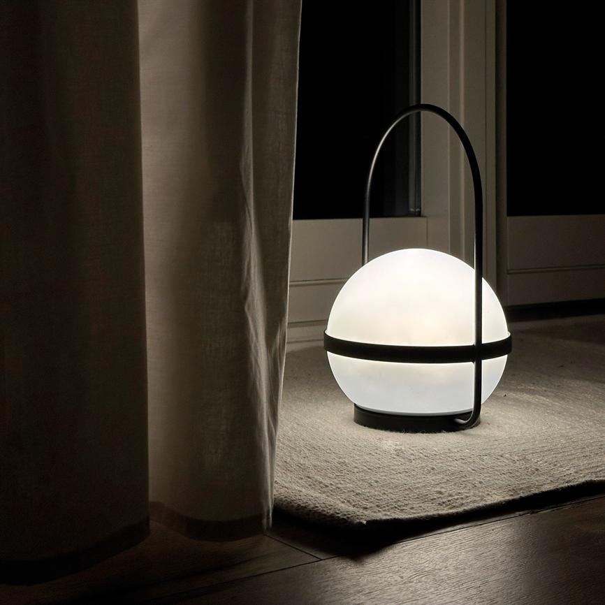 A spherical table lamp with a black bracket emits soft white light and stands on a small rug in front of a closed glass door at night, partially hidden by a curtain.