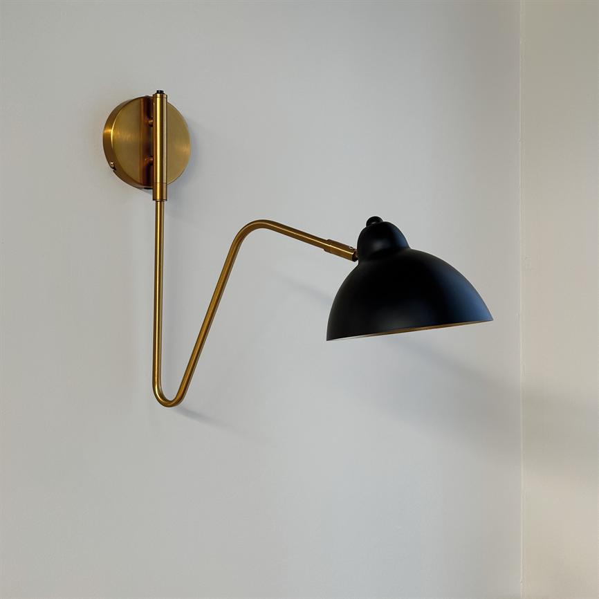 Wall lamp: a brass wall-mounted arm extends in a zigzag and holds a matte black hemispherical shade against a light, neutral wall in a corner.