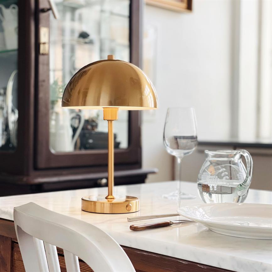 A gold-colored table lamp is lit on a white marble table; casts soft light over a covered space with wine glasses, water carafe, knife and plate; display cabinet and chair in the background.