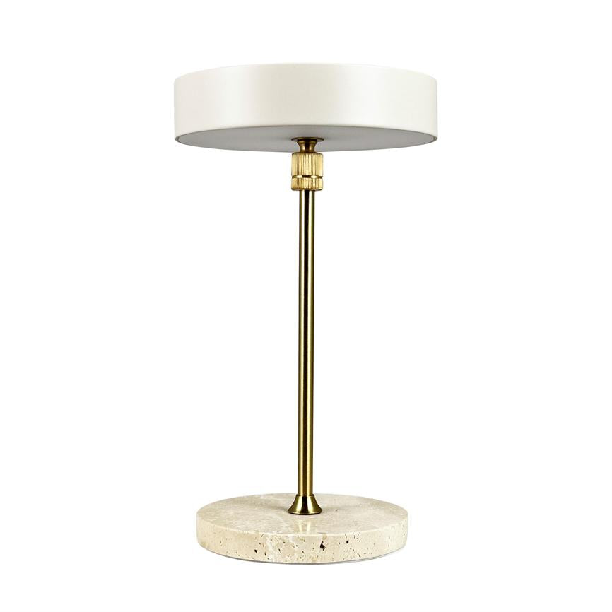Table lamp stands against white background; round, matt white shade, slim brass rod and round travertine foot in light stone.