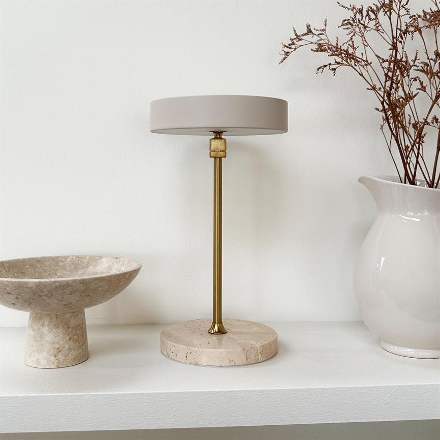 A table lamp stands centrally on a shelf; brass rod and round beige shade rest on marble base, flanked by a stone bowl to the left and a white vase with dry branches to the right.