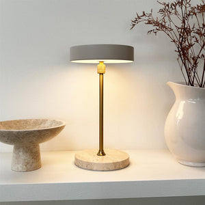 Table lamp stands and lights on a shelf; marble base and brass rod with gray shade. On the left a round marble bowl, on the right a white vase with dried branches.