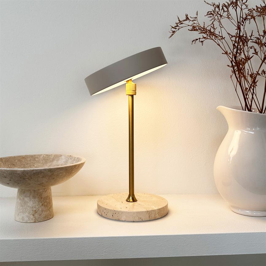 Lamp stands and shines downwards; slender brass rod with round stone base and slanted shade, placed on shelf between a marble bowl and a white jug with dried branches against a light wall.