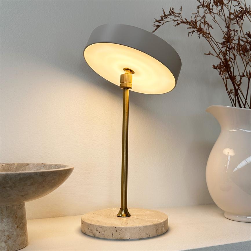 The lamp stands on a round stone base and emits warm light, angled towards the wall; on shelf with a ceramic bowl on the left and a white vase with dried branches on the right.