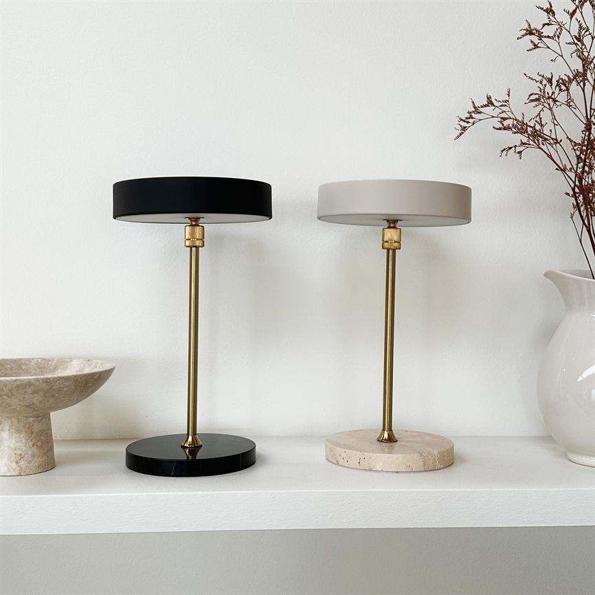 Two table lamps stand on a white shelf; one with black shade and marble base, the other with beige shade and light terrazzo base; golden bars, white wall, vase and bowl.