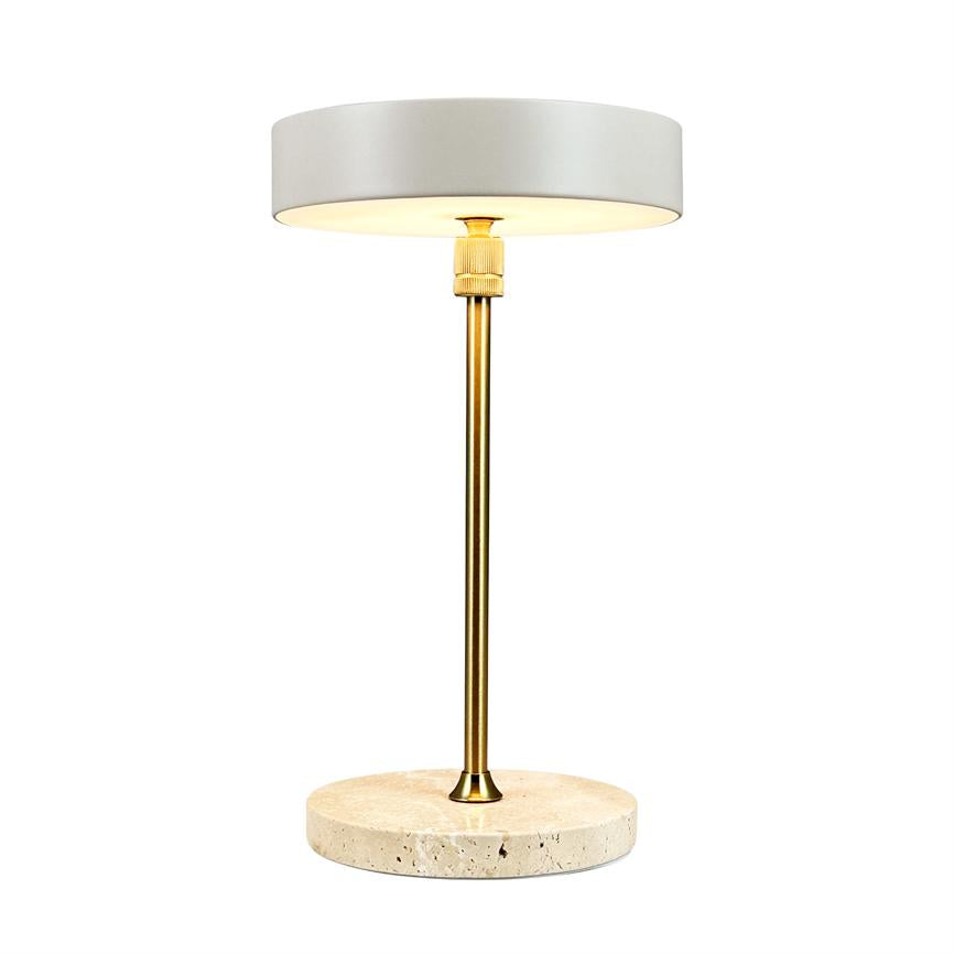 Table lamp is on; white cylindrical shade emits warm light, mounted on a slender brass rod and a round, light travertine base, set against a neutral white background.