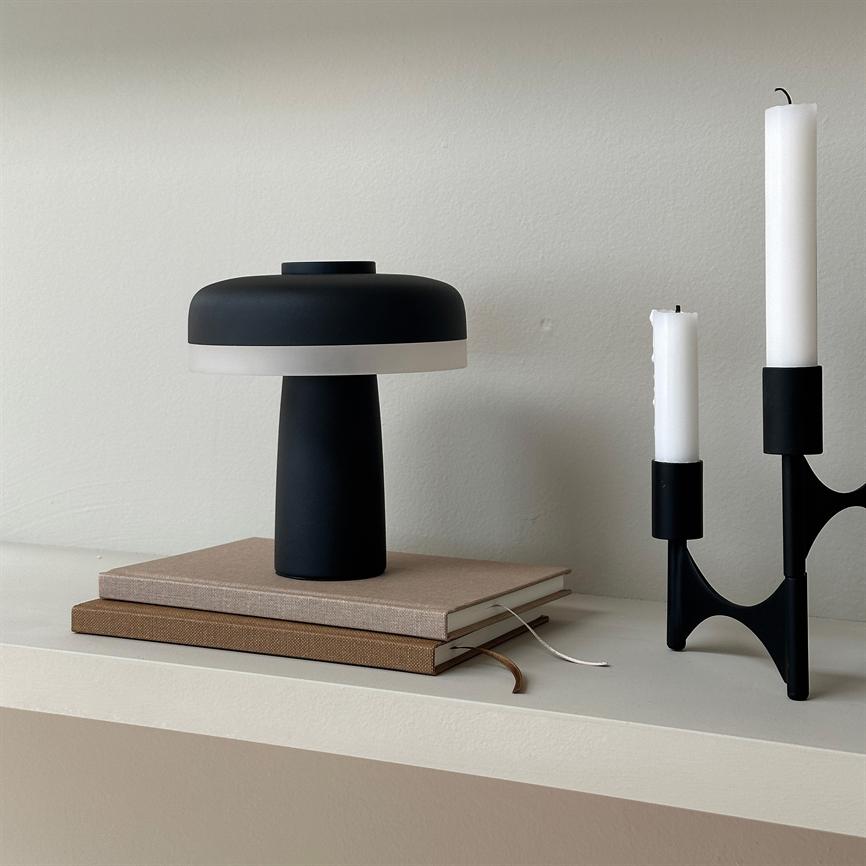 Black table lamp stands on two stacks of notebooks, next to a black candlestick with two extinguished white candles, on a light cream shelf in front of a neutral wall.