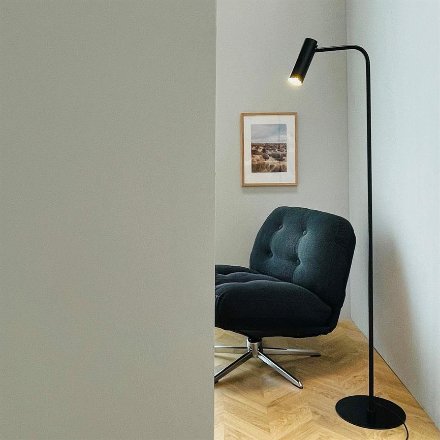 Dark, upholstered swivel chair rests quietly; a black floor lamp casts warm light; a small corner with a light herringbone parquet floor and a framed landscape photo on a light wall.