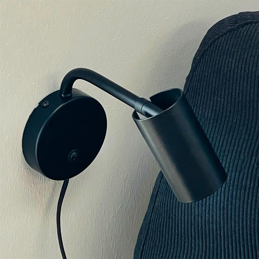 Wall lamp in matt black points downwards; it is mounted on a round wall bracket and hangs over a dark, ribbed fabric back against a light, lightly patterned wall.