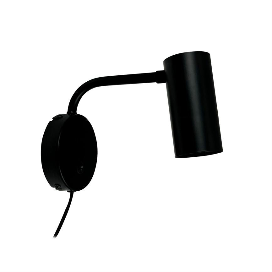 Black metal wall lamp mounted on round wall plate, arm bent outwards and cylinder-shaped lamp head pointing obliquely downwards; simple, modern design against white background.