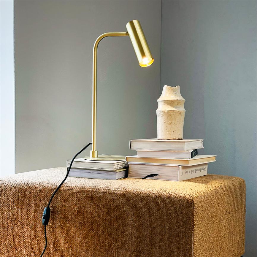 Gold-colored table lamp casts warm rays over a stack of books; a small beige ceramic vase stands on top. The scene is on a brown upholstered pouf in front of a gray wall. No readable text.