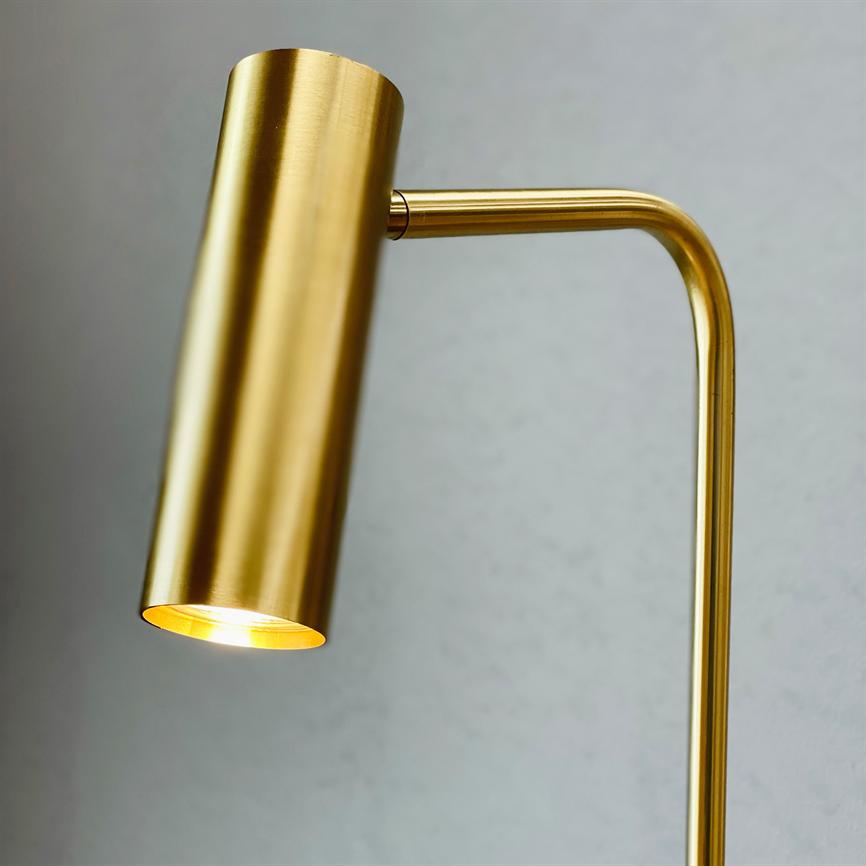 Brass colored table lamp with cylindrical shade, lit and casting warm light downwards; arm bent at a right angle in front of a light gray wall.