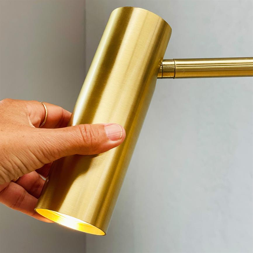 A cylindrical, brass-plated lamp is held and adjusted by one hand; light shines from the opening of the lamp, mounted on a thin arm in front of a light gray wall.