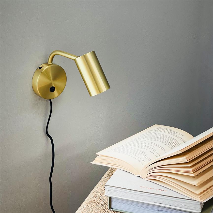 Gold-colored wall lamp points down and illuminates an open book lying on a stack of books on a bedside table; the lamp is mounted on a gray wall with a visible black cord. There is printed text in the book, but it is not clearly legible.